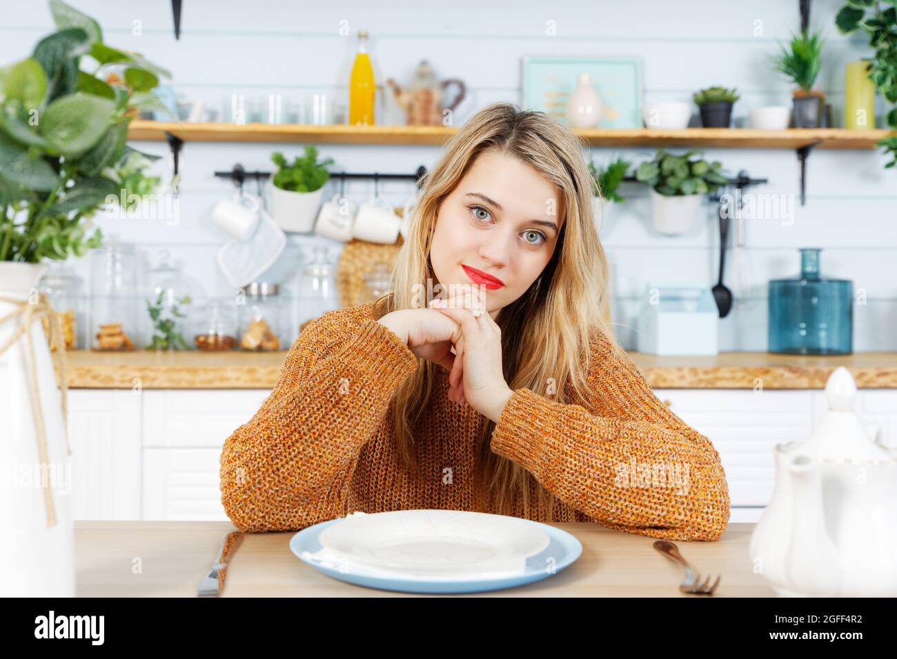 Jeune femme assise à la table de cuisine devant une assiette vide. Arrière-plan flou. Banque D'Images Jeune femme assise à la table de cuisine devant une assiette vide. Arrière-plan flou. Banque D'Images