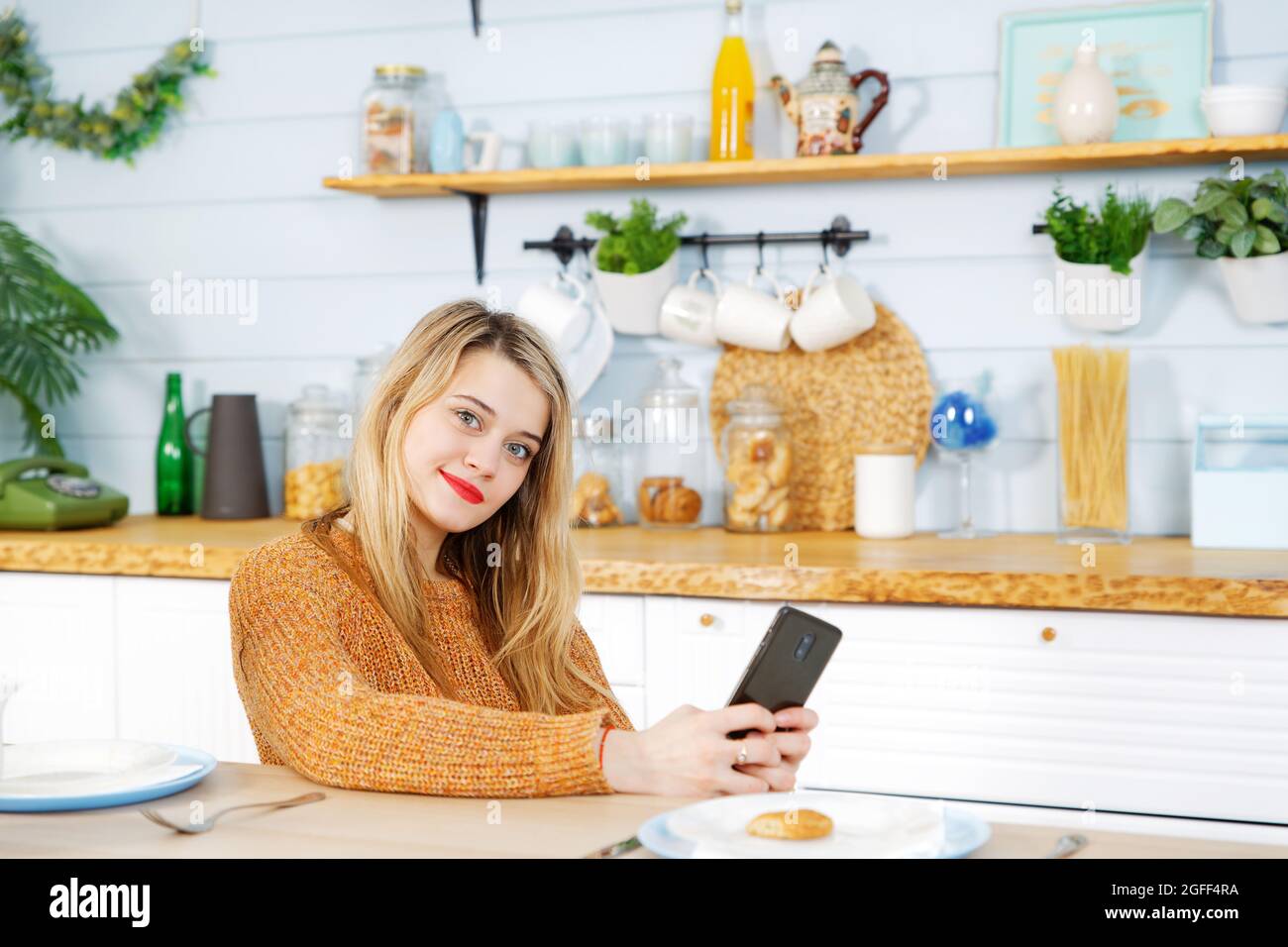 Jeune jolie fille assise à une table dans la cuisine avec un téléphone dans ses mains Banque D'Images Jeune jolie fille assise à une table dans la cuisine avec un téléphone dans ses mains Banque D'Images