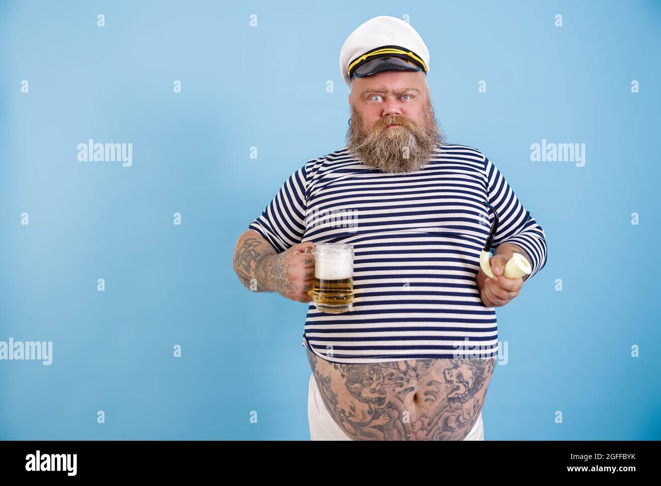 Drôle de marin obèse tient fumer le tuyau et la bière grimacing sur fond bleu clair Banque D'Images