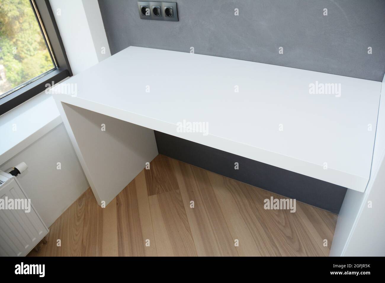 Une table d'ordinateur au fini blanc brillant sur le plancher en bois stratifié près de la fenêtre avec radiateur et mur gris peint avec multiple de montage mural européen noir Banque D'Images