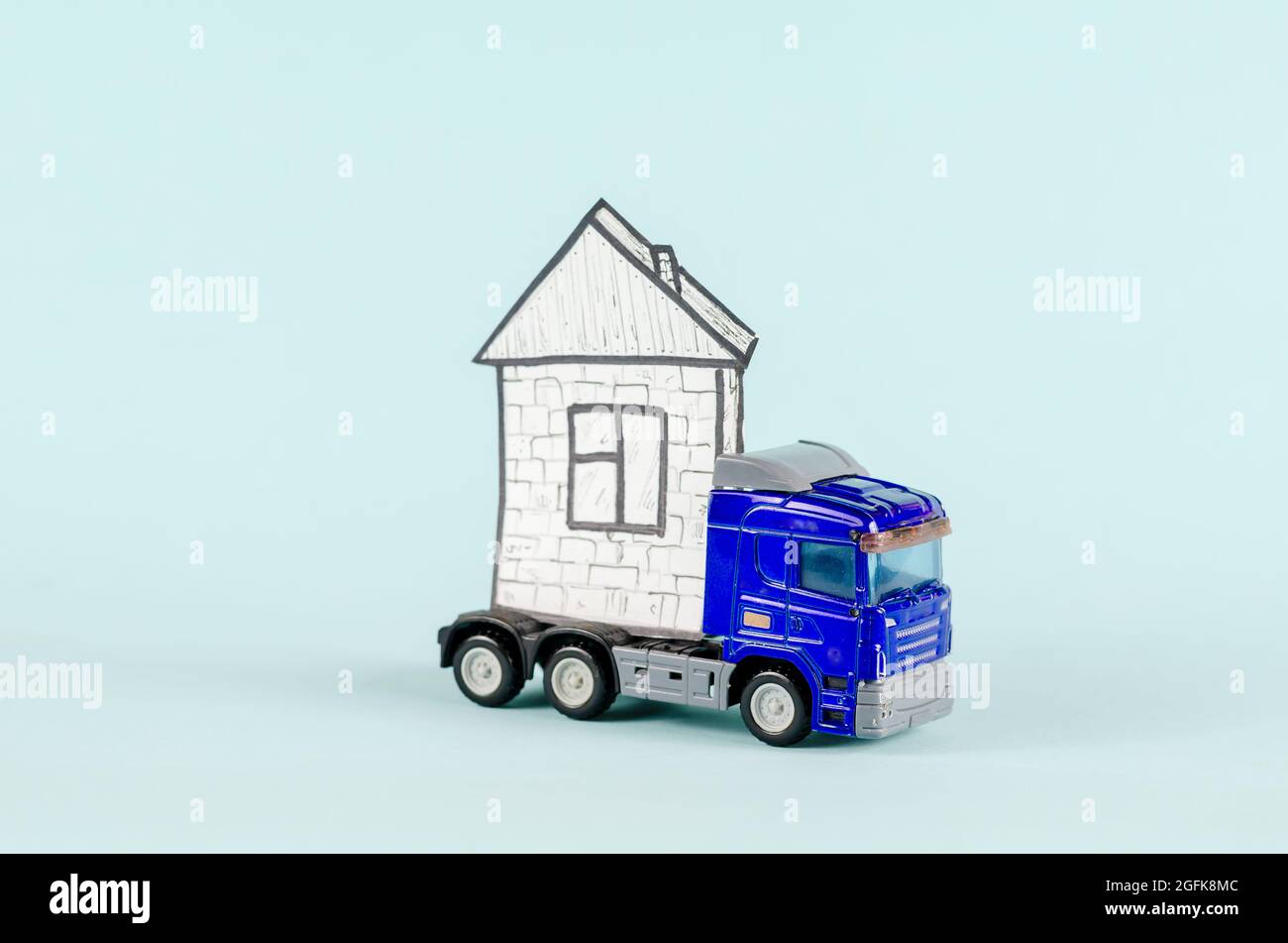 Tracteur-remorque jouet transportant une maison. Chariot en plastique bleu et dessin de maison sur papier blanc. Arrière-plan bleu. Immobilier, industrie, transports. Sél Banque D'Images