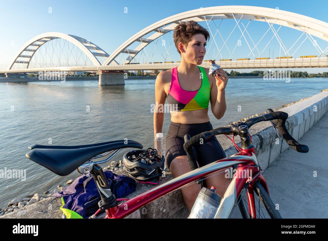 Bonne jeune femme cycliste mangeant une barre d'énergie, rafraîchissant et  ayant un repos au bord de la rivière, après avoir porté un vélo. Un mode de  vie sain et des sports Photo