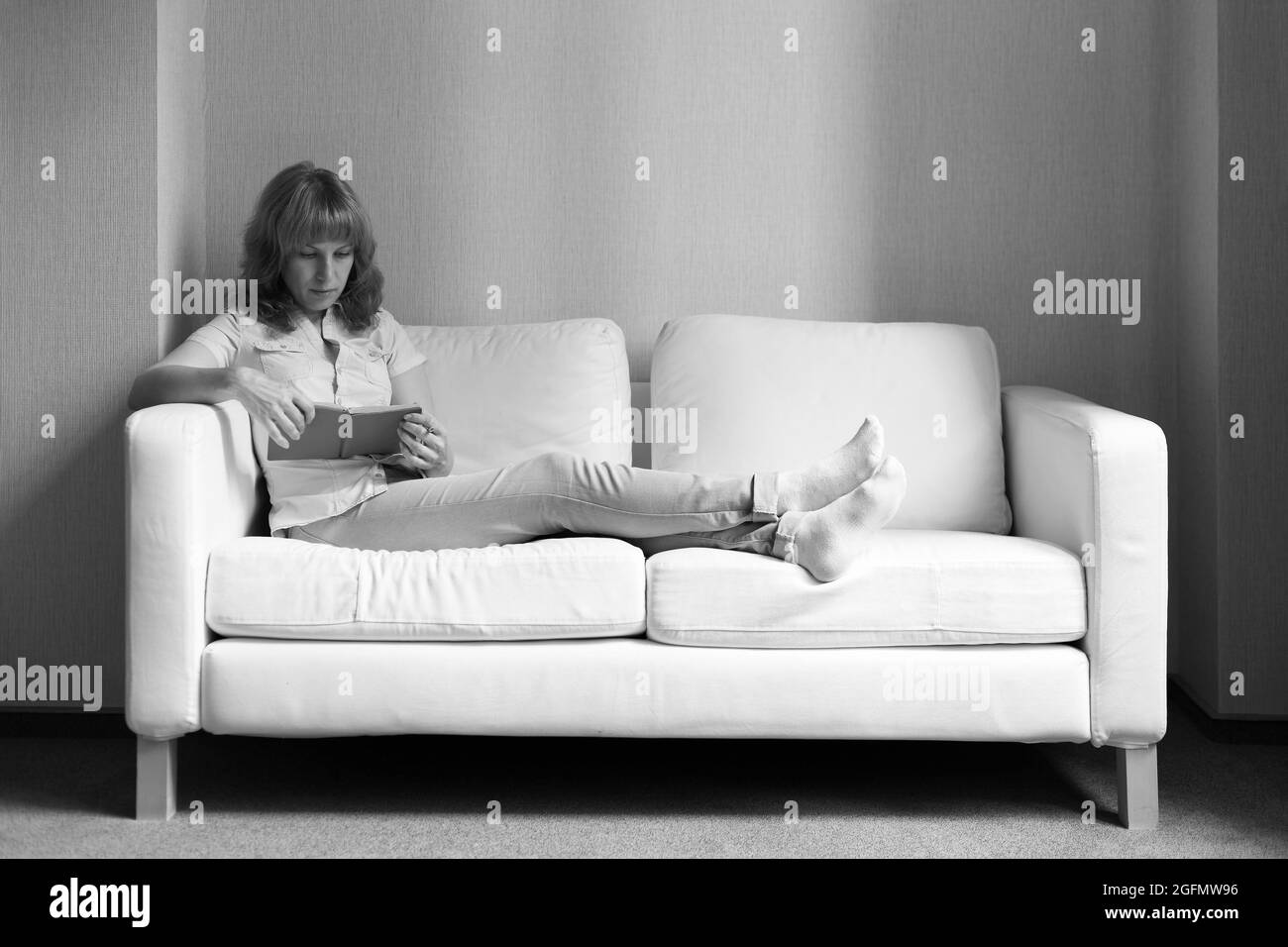 Une jeune femme dans une chemise bleu clair et un Jean assis sur un canapé et tenant un livre dans la main. Image en noir et blanc. Banque D'Images