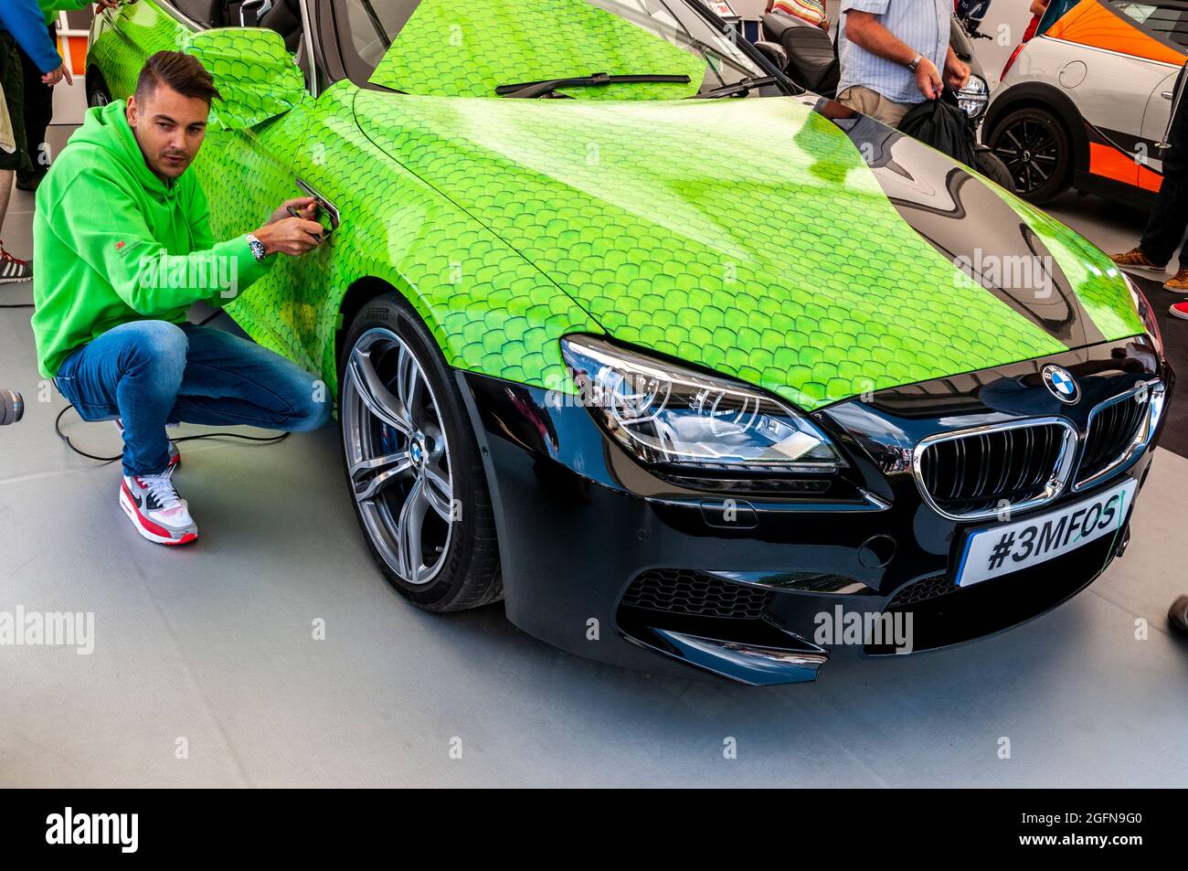 Un homme démontrant l'emballage d'une voiture BMW dans une finition de style écailles de poisson vertes. Démonstration professionnelle 3M de la couverture adhésive sur mesure Banque D'Images