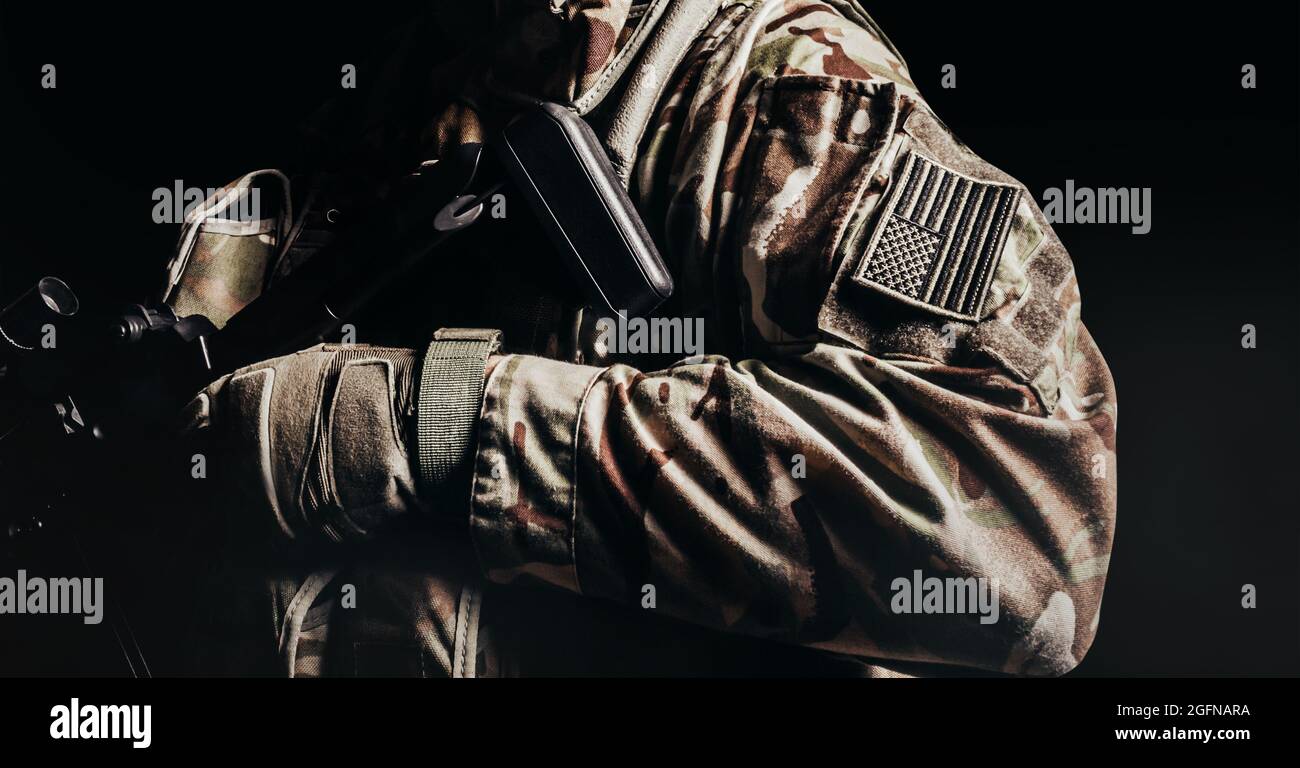 Photo d'un soldat américain ombré en gants de protection uniformes et tactiques avec étiquette drapeau américain tenant le fusil sur fond noir. Banque D'Images
