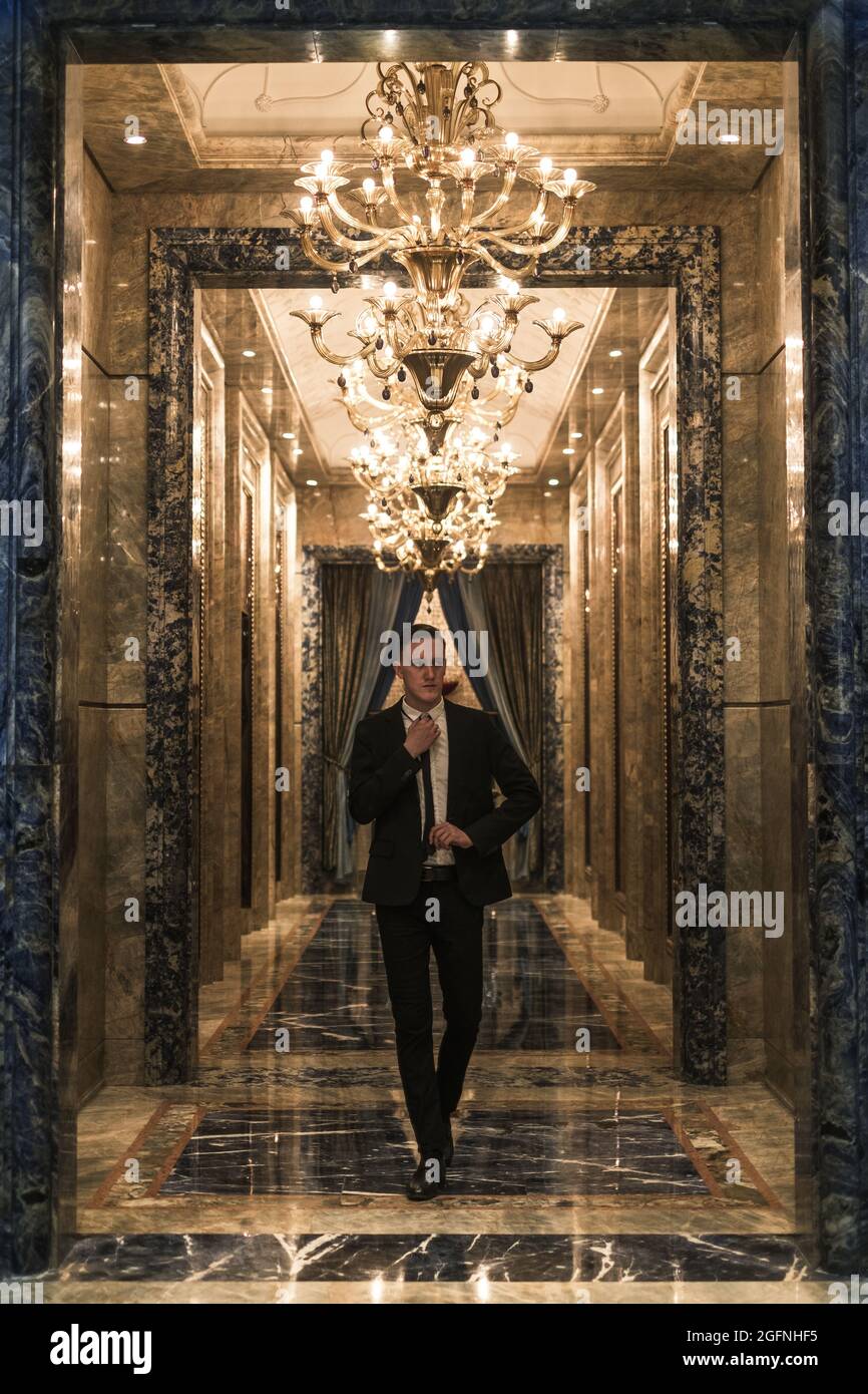 Jeune homme d'affaires caucasien élégant en costume d'affaires marchant dans le hall avec de beaux lustres chers. Redresse sa cravate Banque D'Images