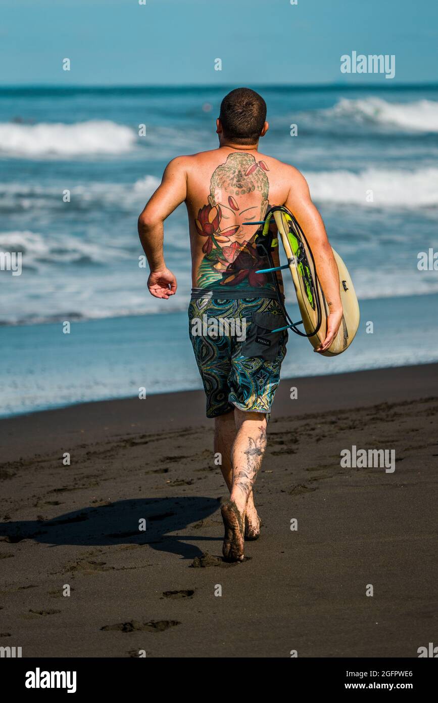 Playa Hermosa, Guanacaste, Costa Rica - 07.26.2020: Un homme bien construit avec des tatouages colorés porte une planche de surf et court vers l'océan. Banque D'Images
