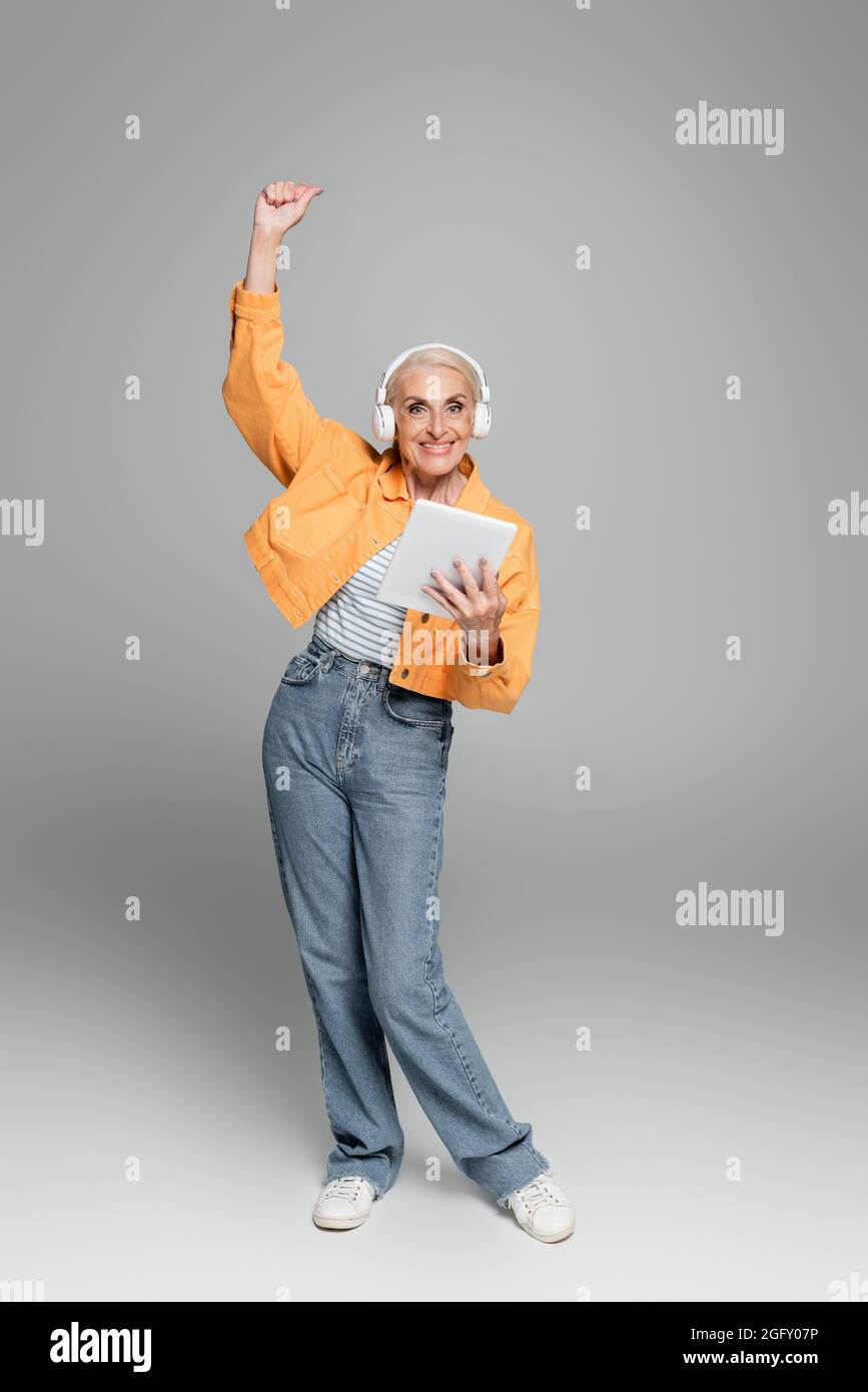 femme âgée joyeuse dans un casque tenant une tablette numérique et montrant le geste de gain sur le gris Banque D'Images
