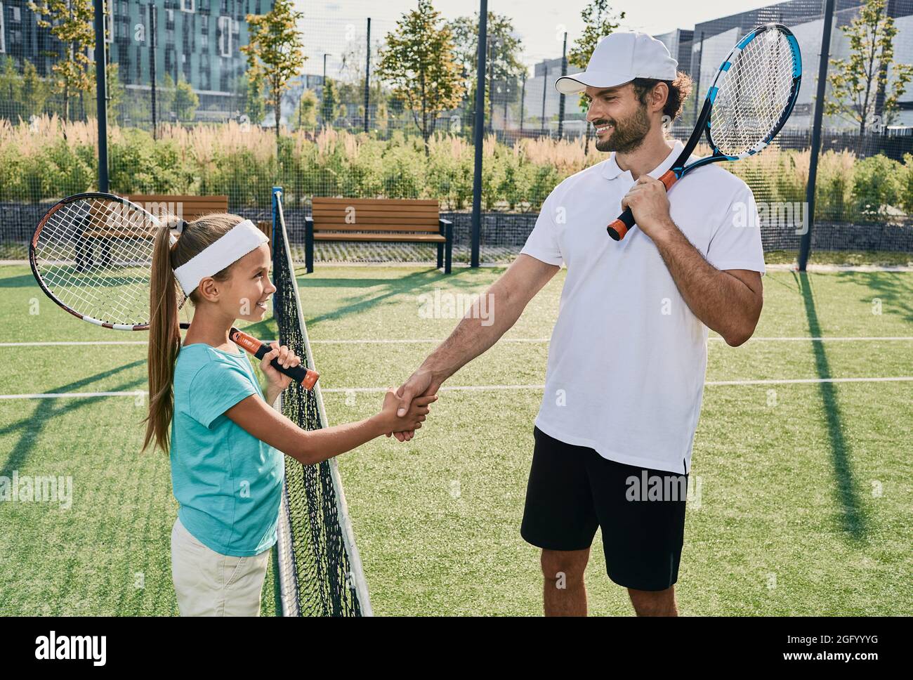 Une petite fille secoue les mains avec son entraîneur professionnel près du filet de tennis avant de jouer au tennis. Apprentissage du tennis pour les enfants Banque D'Images