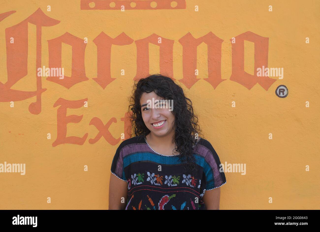 Belle jeune femme mexicaine Latina avec de longues boucles sourires devant un mur de maison jaune avec peint Corona Extra bière publicité. Banque D'Images