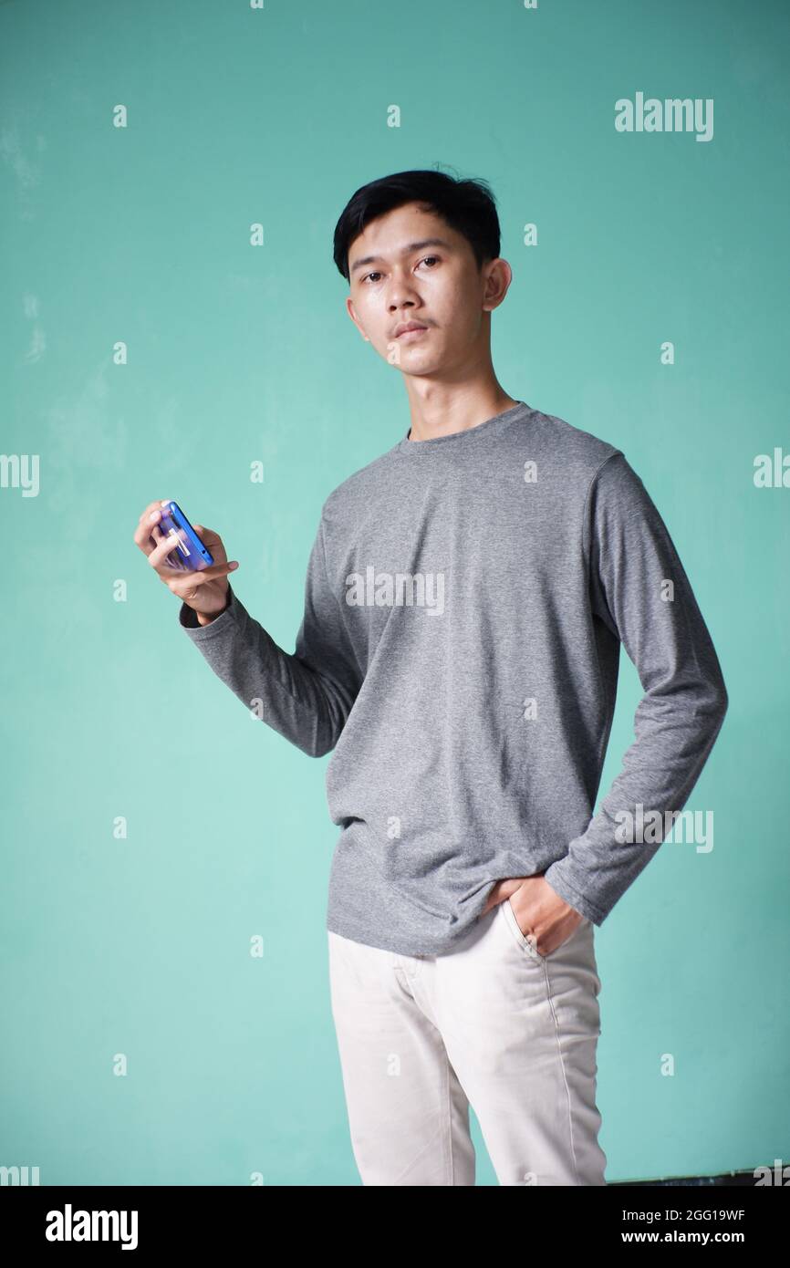 portrait d'un homme asiatique beau portant un t-shirt gris avec une posture fraîche. homme moulant sur fond vert. homme tenant un téléphone portable avec une expression sérieuse Banque D'Images