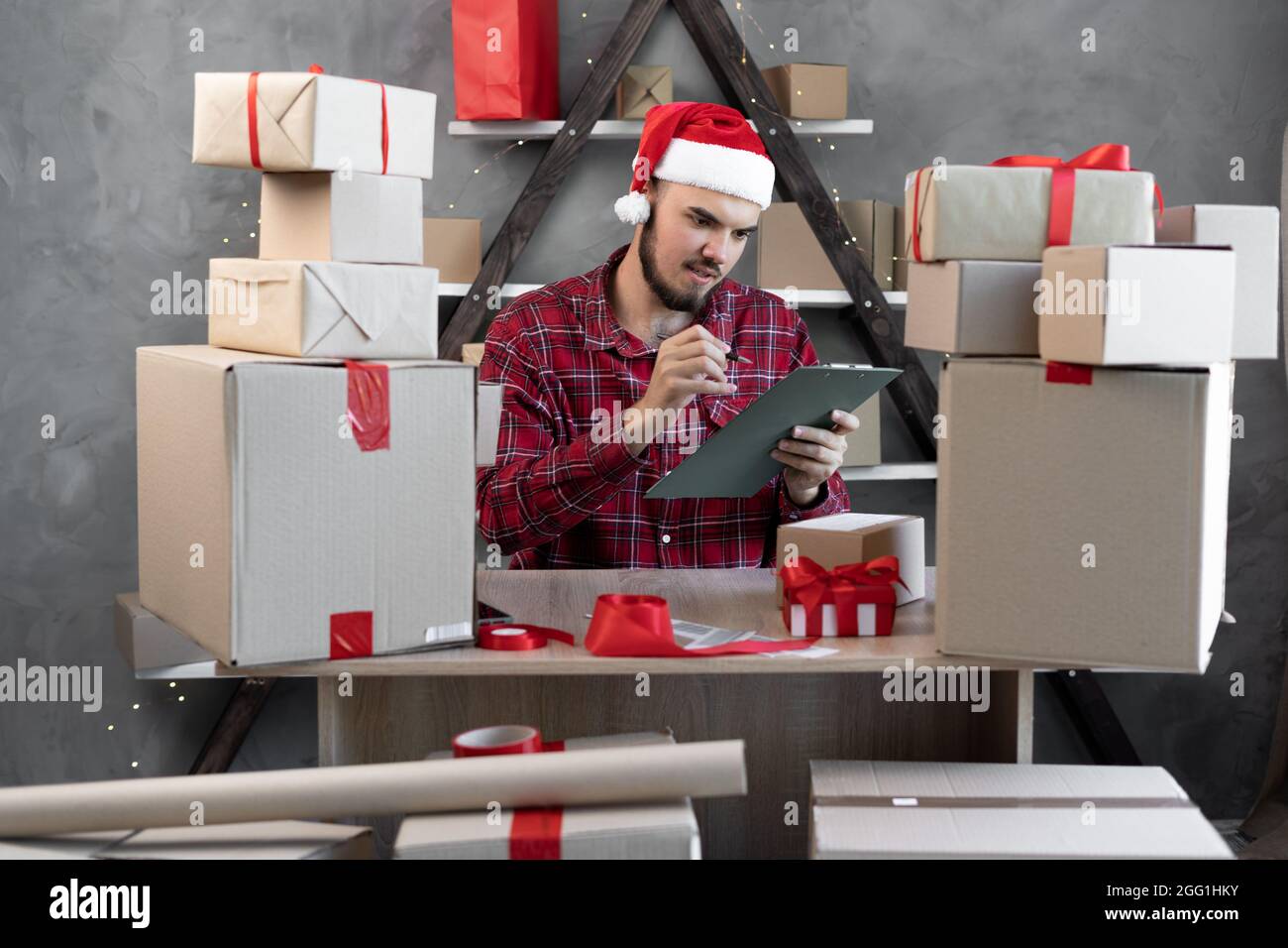Le propriétaire de petite entreprise Santa Claus écrit une liste d'acheteurs ou de clients travaillant dans un entrepôt ou un atelier à la veille de Joyeux Noël à côté des boîtes o Banque D'Images