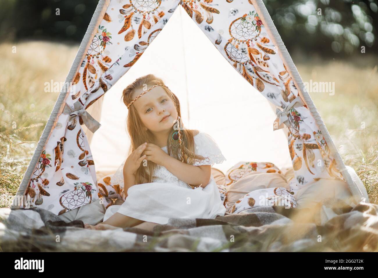 Adorable petite fille blonde assise dans une tente de tipi de boho.  Adorable fille enfant, portant la robe blanche et les accessoires de  cheveux de plumes dans le style natice américain, posant