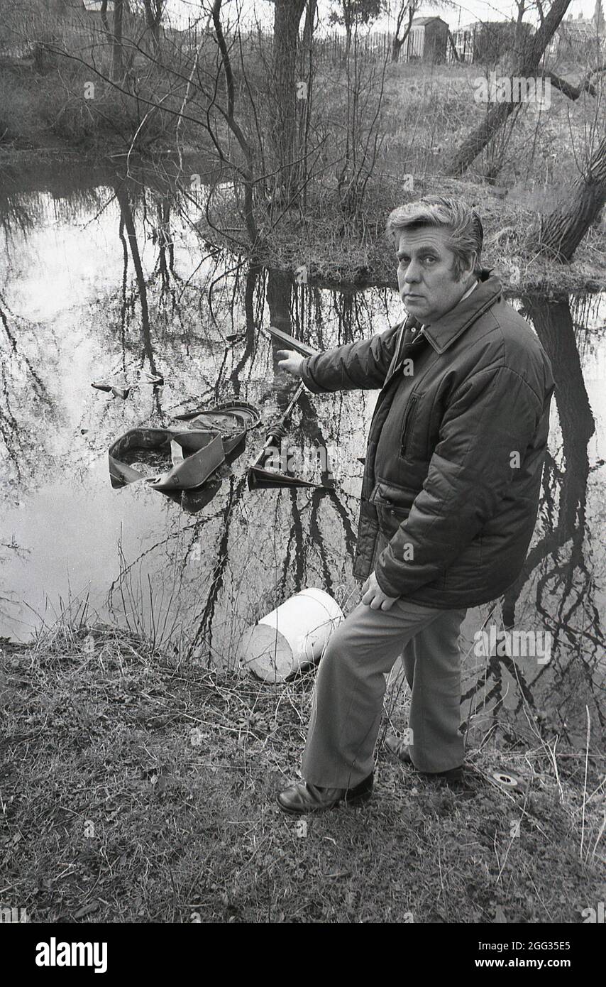 1987, historique, un homme dans un anorak extérieur pointant vers les articles domestiques jetés, y compris une vieille valise, qui ont été jetés dans un étang de bois, Angleterre, Royaume-Uni. Banque D'Images