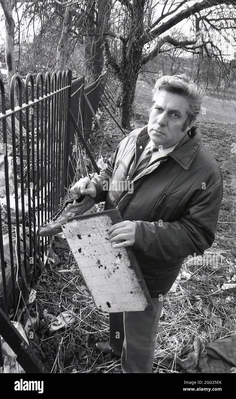 1987, historique, un homme dans un anorak tenant jeté des articles domestiques, une chaussure de sport et de plancher, qui ont été laissés ou jetés à côté d'une clôture de parc, Angleterre, Royaume-Uni. Banque D'Images