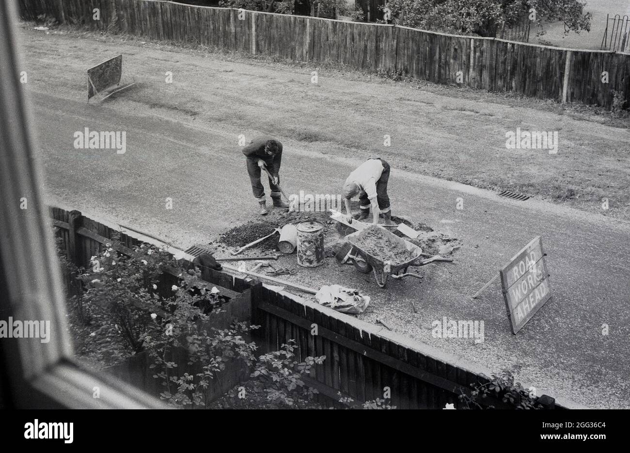 Années 1960, historique, vue d'une fenêtre à l'étage de deux ouvriers à l'extérieur faisant des réparations sur une route de village, Witney, Oxford, Angleterre, Royaume-Uni. Banque D'Images