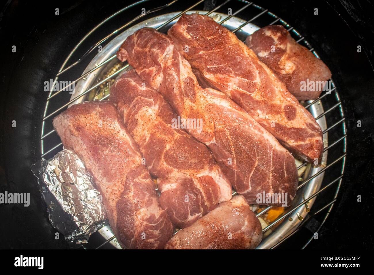 Côtelettes de porc assaisonnées crues sur le gril à l'intérieur de la nourriture cylindrique fumeur avec des herbes dans le papier d'aluminium à côté Banque D'Images