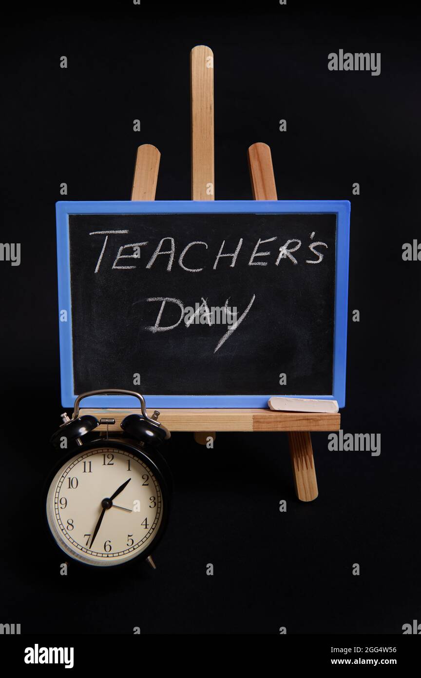 Gros plan d'un tableau noir à craie avec lettrage pour la Journée de  l'enseignant, debout sur un chevalet de table en bois, à côté d'un réveil  noir, isolé sur fond noir Photo