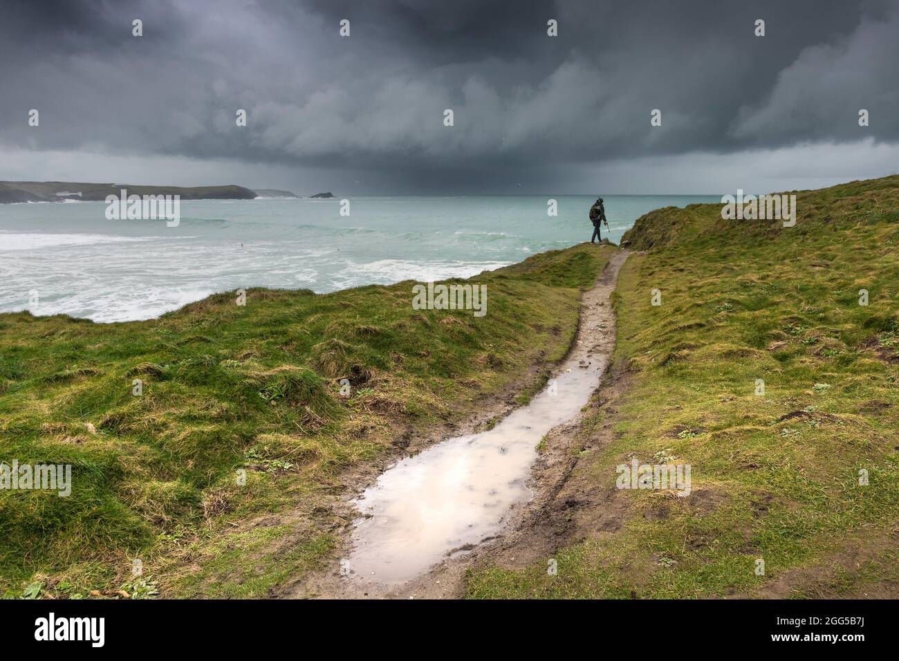 Un pêcheur à la ligne marchant le long du sentier côtier avec des nuages de tempête spectaculaires approchant de la baie de Fistral à Newquay, dans les Cornouailles. Banque D'Images