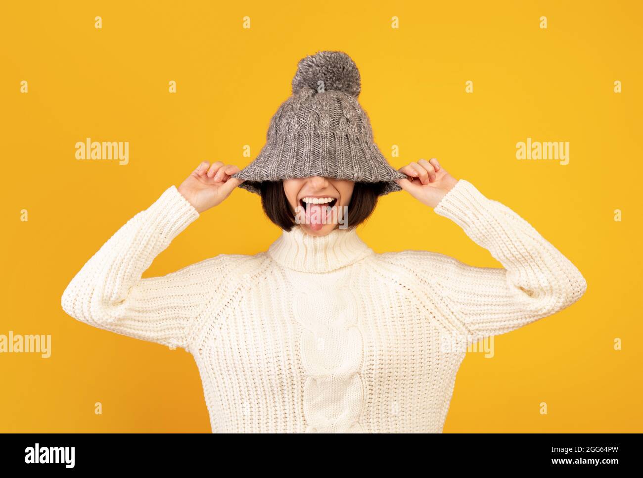 Jeune femme ludique tirant vers le bas chapeau de laine et en collant hors de sa langue, se moquant sur fond jaune Banque D'Images