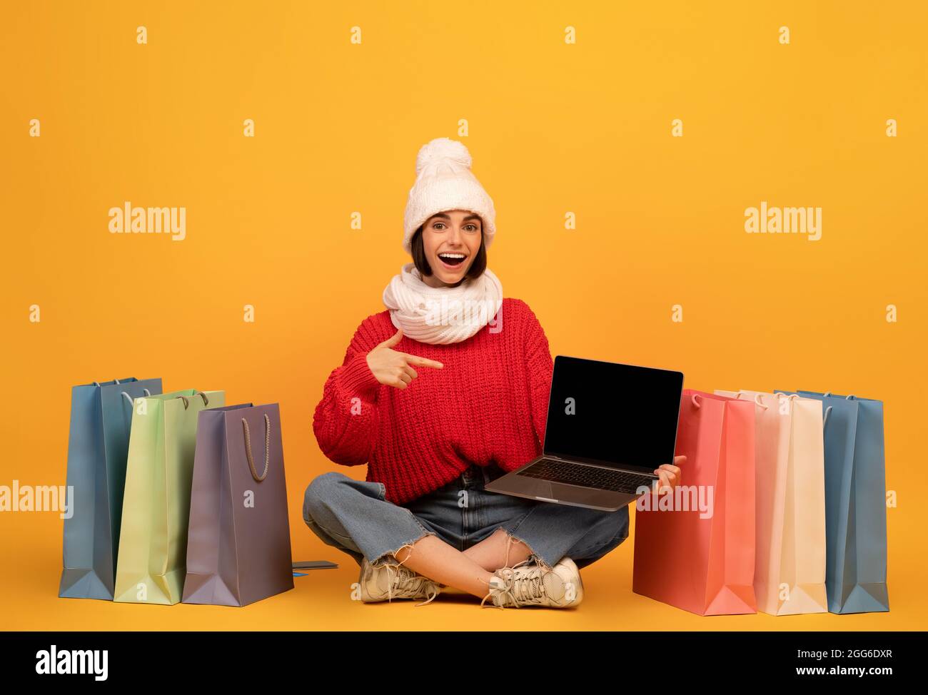Bonne femme shopaholic en tenue d'hiver pointant à l'écran d'ordinateur portable, assis parmi les sacs de cabas, fond jaune Banque D'Images
