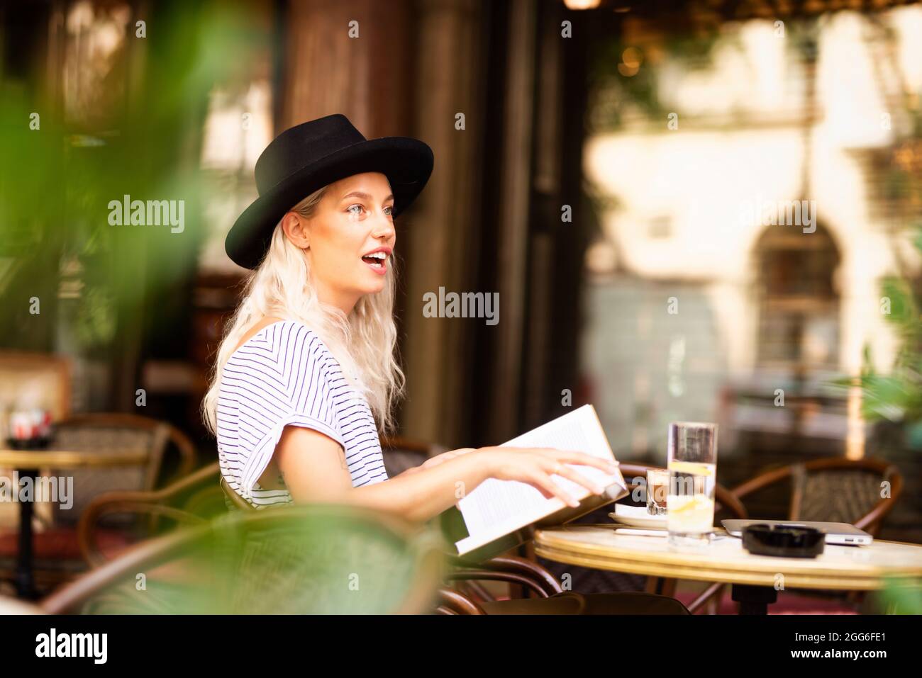 Petit cliché de jeune femme blonde aux cheveux portant un chapeau fedora et  lisant un livre tout en discutant avec quelqu'un sur le café extérieur  Photo Stock - Alamy