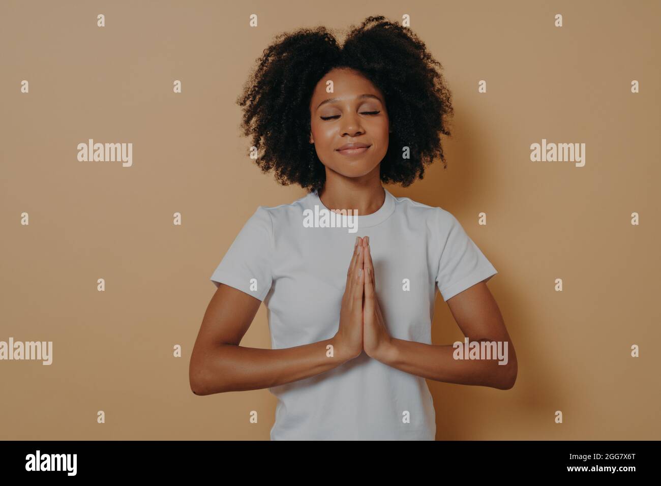 Une jeune femme afro-américaine tenant les mains jointes et priant sur fond beige Banque D'Images
