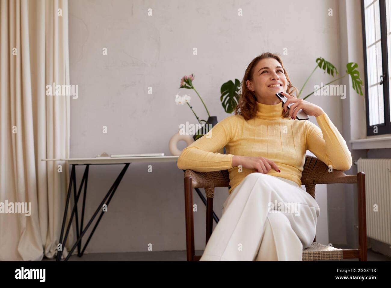 Jeune femme vêque de vêtements décontractés, tenant un téléphone portable et rêvant volontiers tout en étant assise sur une chaise à la maison Banque D'Images