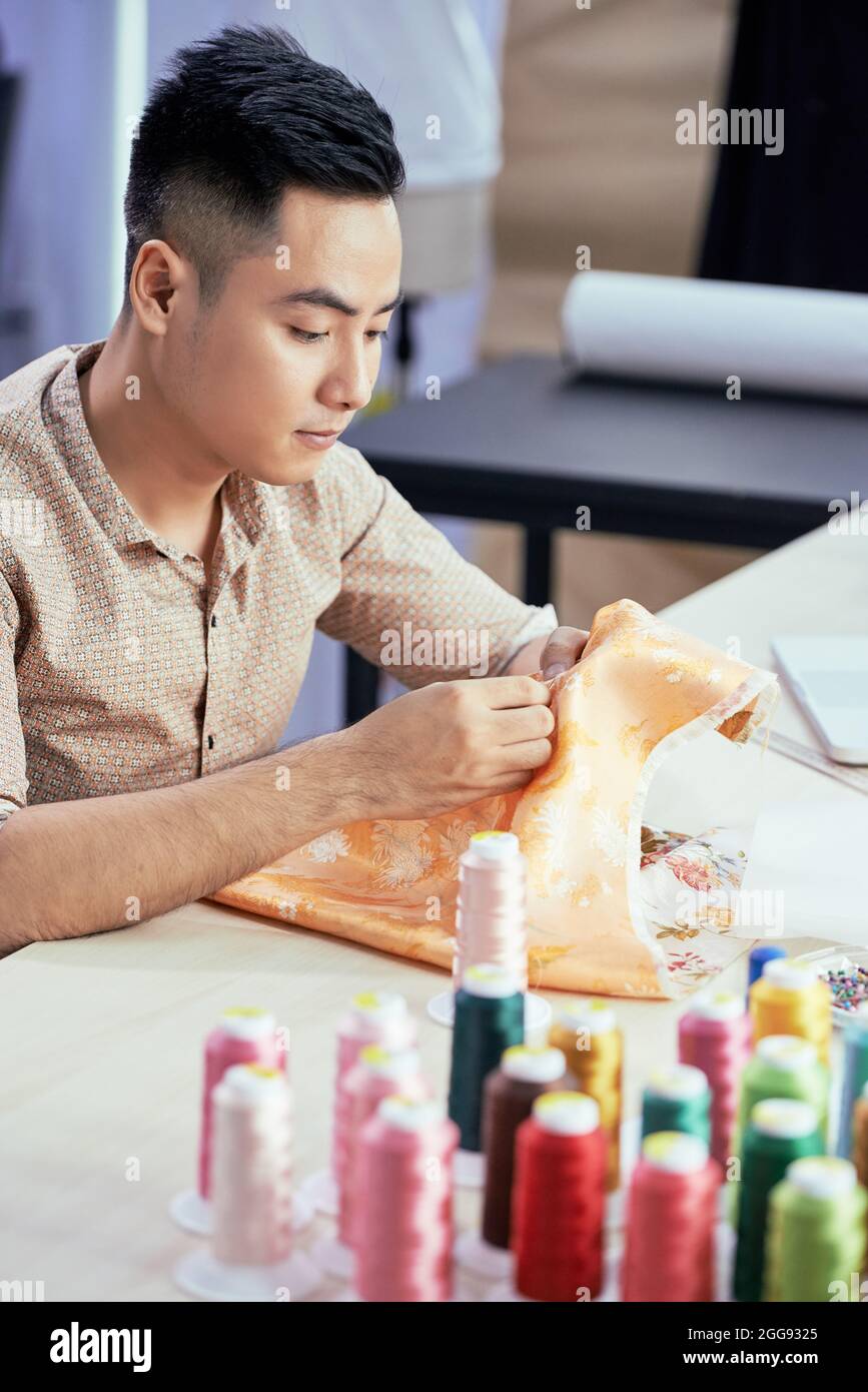 Jeune homme créatif talentueux tailleur travaillant avec du tissu en soie coûteux à sa table dans l'atelier Banque D'Images