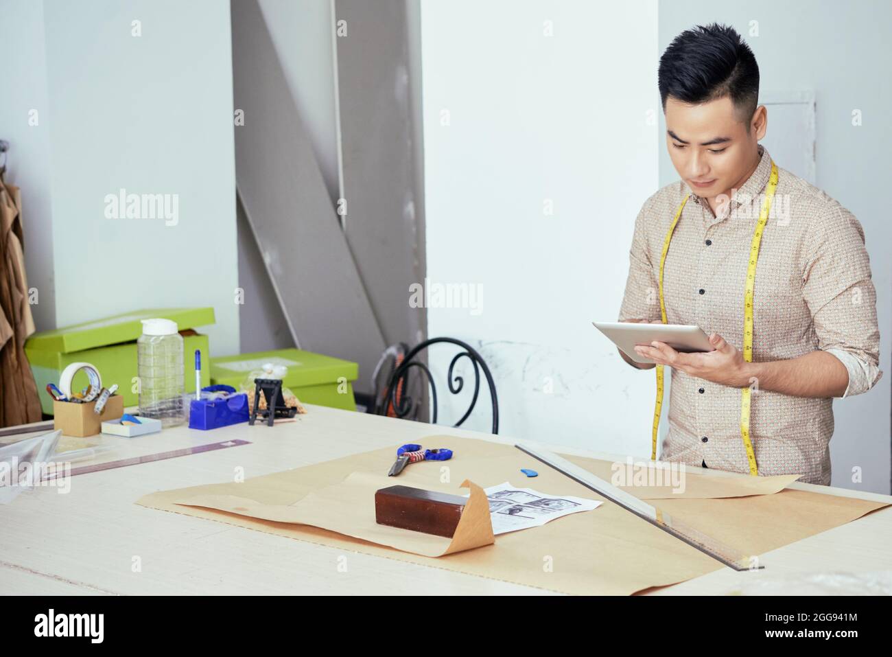 Jeune homme tailleur vérifiant ses notes sur l'ordinateur tablette lors de dessin de modèle de couture sur la grande feuille de papier Banque D'Images