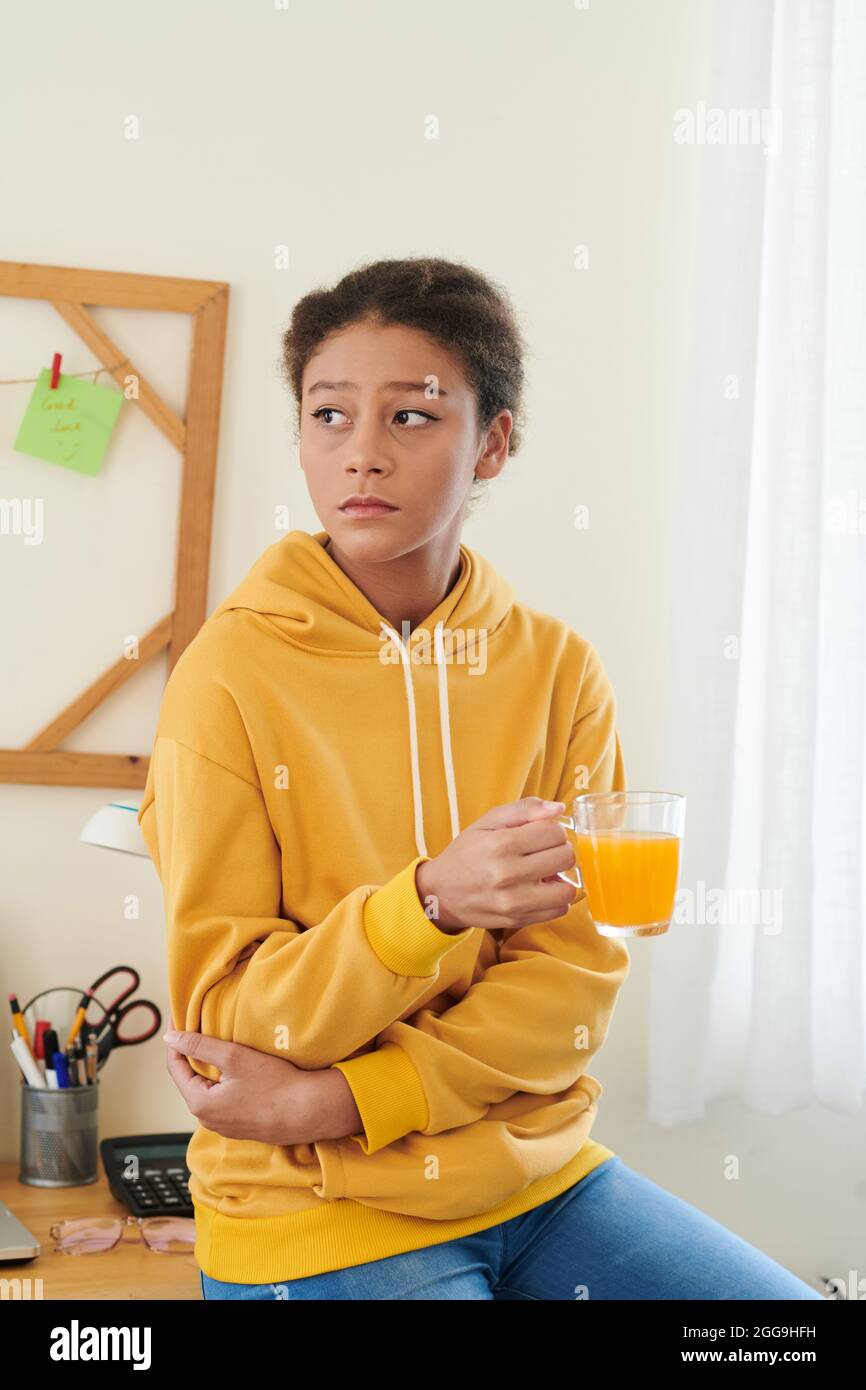 Triste adolescente fatiguée à capuchon jaune assise sur son bureau à la maison et buvant une tasse de jus d'orange Banque D'Images