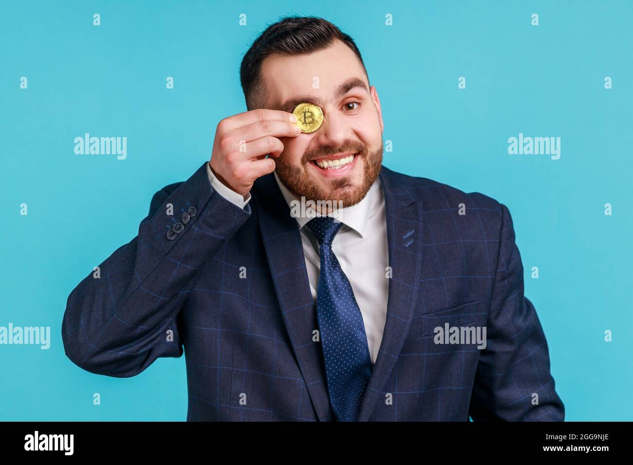 Portrait d'un homme d'affaires heureux portant un costume de style officiel foncé couvrant l'œil avec du bitcoin doré, regardant l'appareil photo à travers la pièce de btc. Studio intérieur sho Banque D'Images