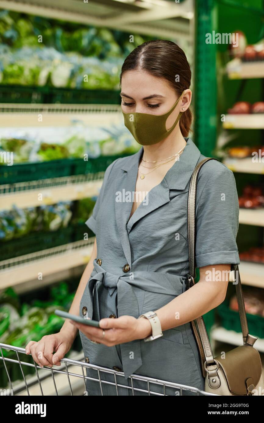 Jeune femme enceinte en masque facial et robe debout au panier dans le département des légumes du supermarché et liste de contrôle sur le téléphone Banque D'Images