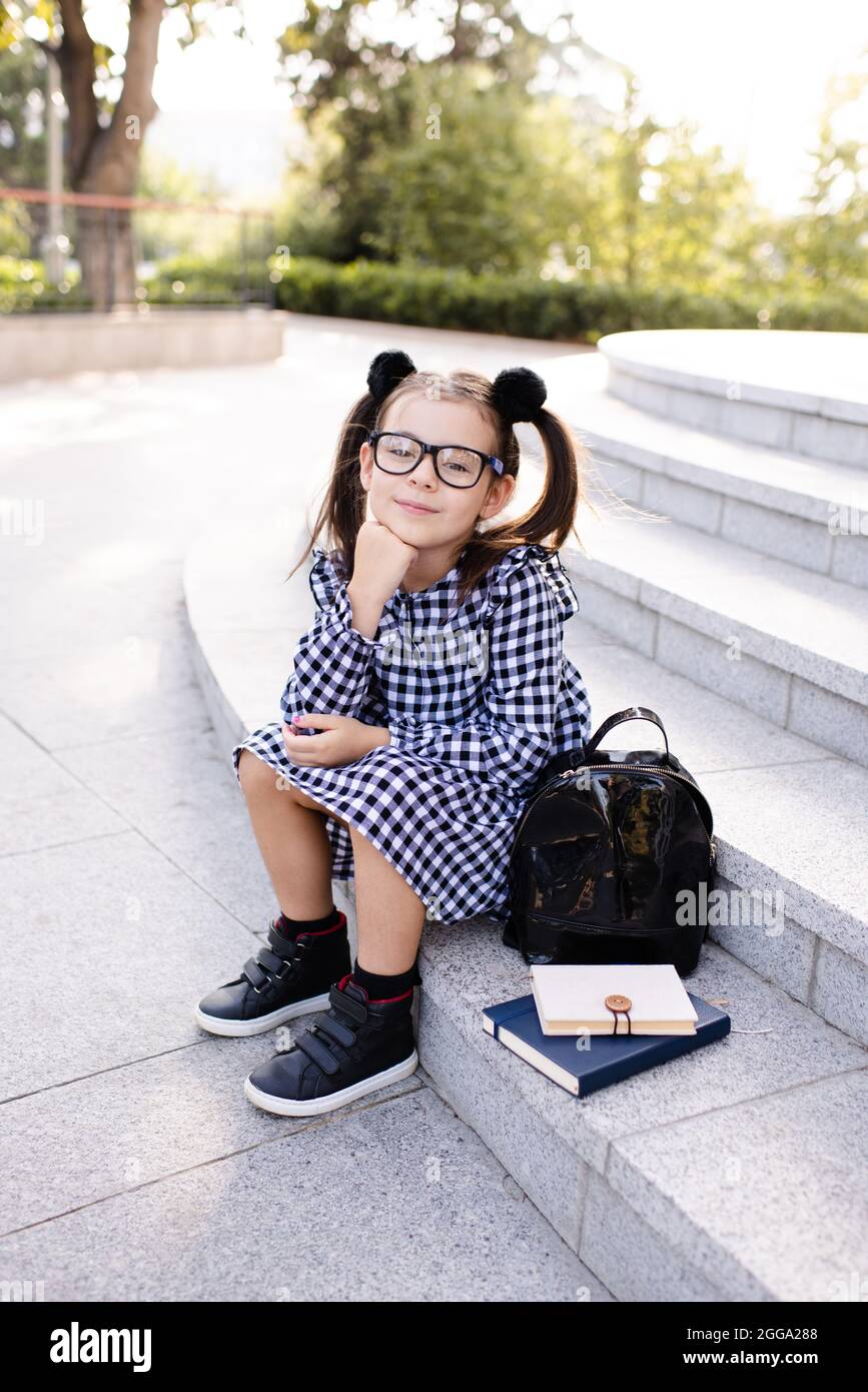 Sourire heureux enfant fille 5-6 ans porter uniforme robe et des lunettes assis sur l'escalier avec des livres et sac à dos dehors. Regarder l'appareil photo. Retour à la goélette Banque D'Images