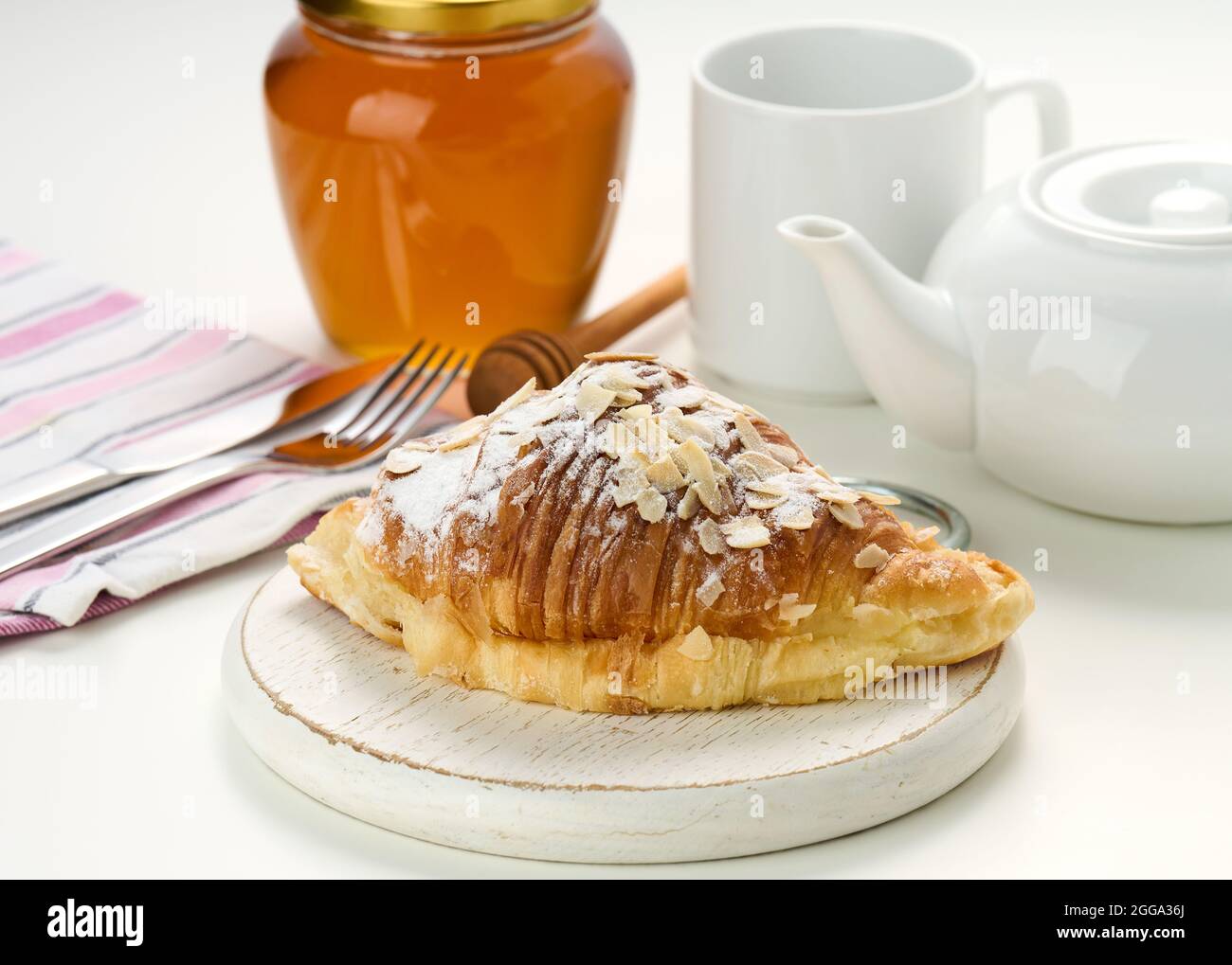 Un croissant croustillant cuit est arrosé de sucre en poudre et de flocons d'amandes sur un panneau de bois, une infusion de céramique blanche et une tasse sur une table blanche. Petit déjeuner Banque D'Images