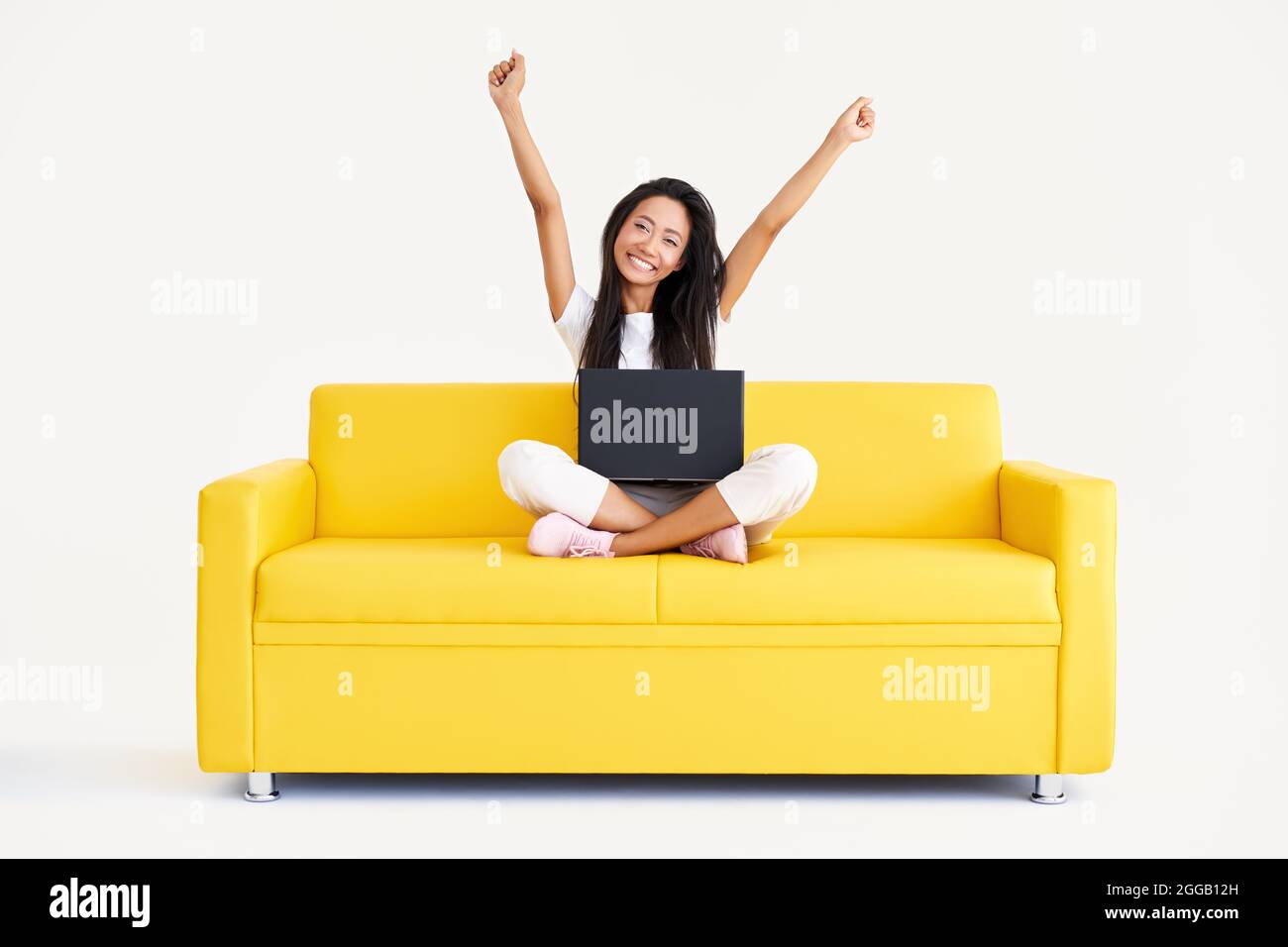 Femme souriante et heureuse avec bras relevés assise sur un canapé jaune avec ordinateur portable et regardant la caméra sur fond blanc. Victoire, célébration, succès conc Banque D'Images