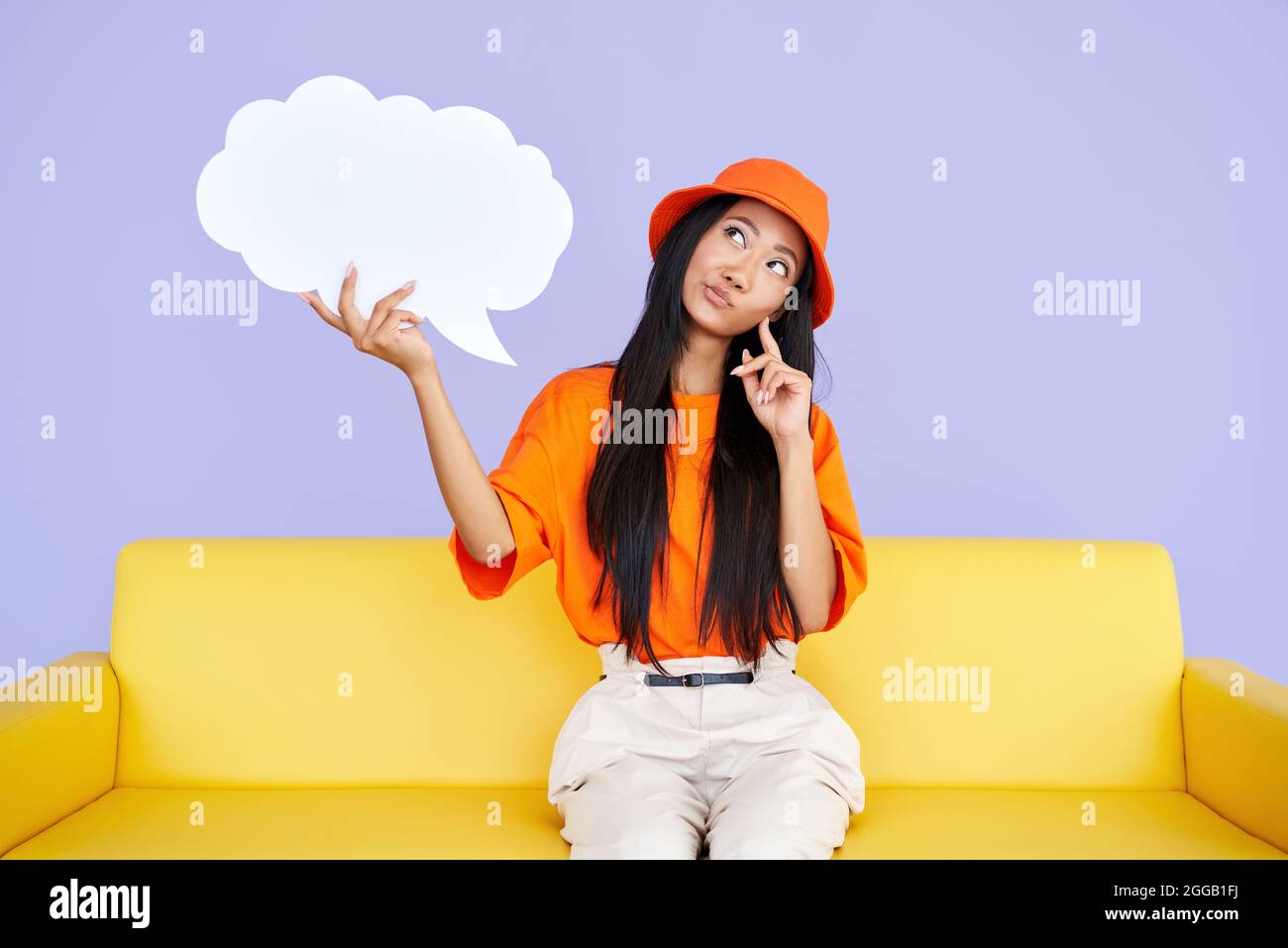 Femme hipster pensive tenant une bulle vide de parole regardant vers le haut assis sur un canapé jaune. Banque D'Images