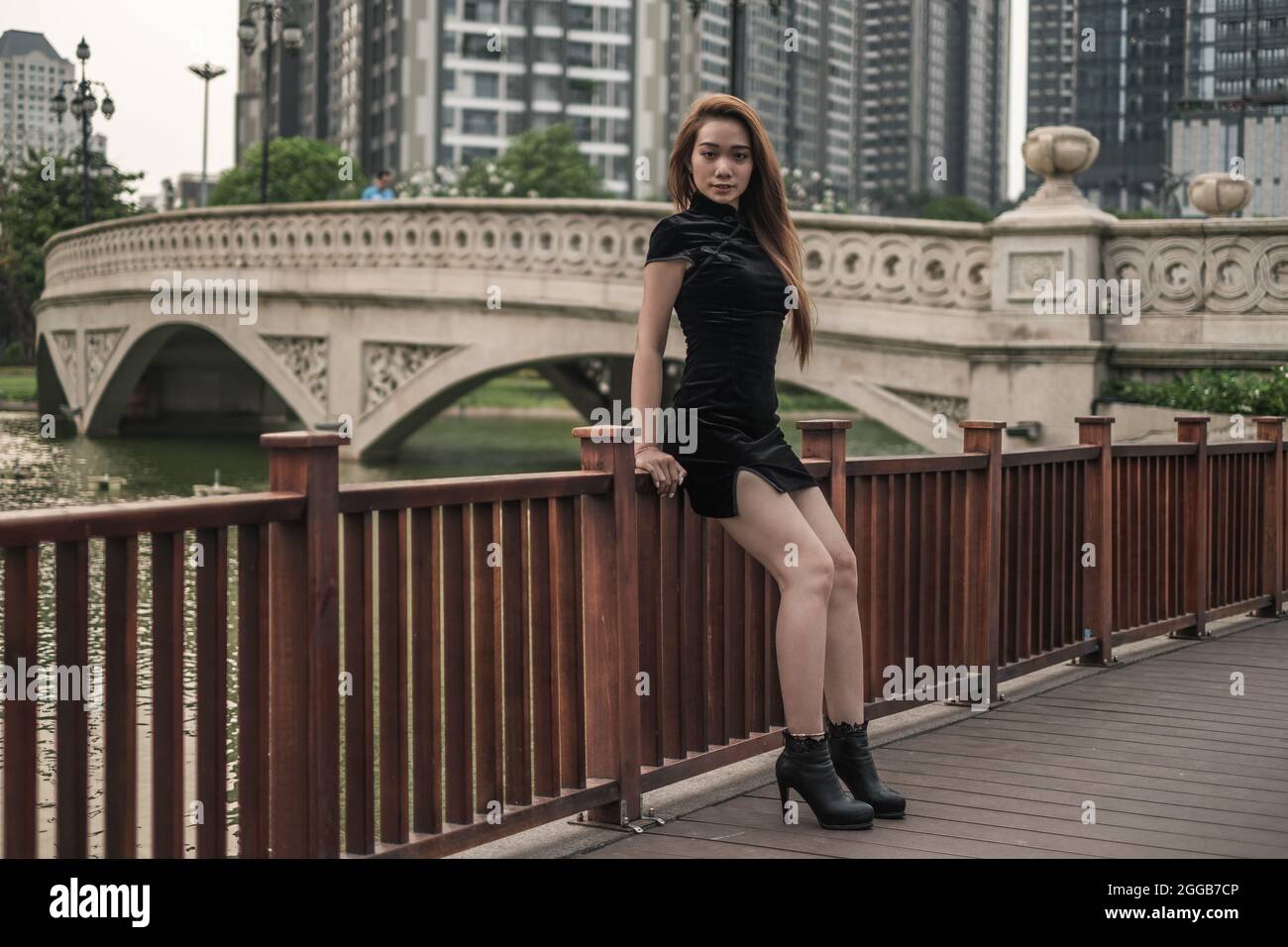 Belle jeune femme asiatique en robe noire mini debout près de la main courante dans le parc. Regarder l'appareil photo avec l'espace de copie. Penché sur la main courante Banque D'Images