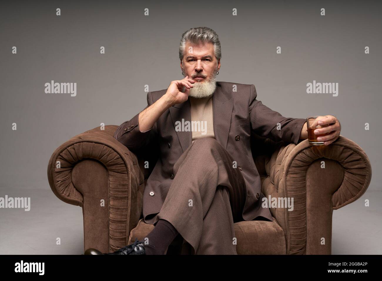 Homme d'affaires mûr et attentionné avec barbe vêtue d'un costume élégant regardant l'appareil photo, tenant un verre de whisky assis dans un fauteuil sur un gris clair Banque D'Images