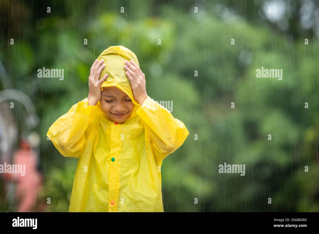 Un garçon portant un imperméable jaune dans un parc public. Joyeux petit enfant asiatique s'amusant à jouer avec les gouttes de pluie. Un garçon qui profite de la pluie. Banque D'Images