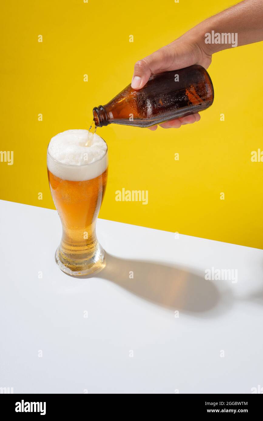 Un bras sert une bière dans un verre, avec de la mousse blanche, sur une table blanche et un fond jaune Banque D'Images