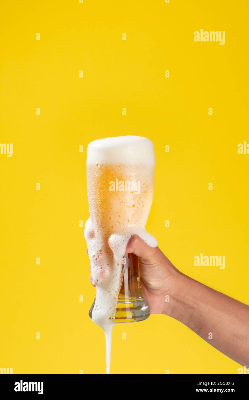 le bras de l'homme tient un bécher en verre avec de la bière à l'intérieur, de la mousse blanche et un fond jaune uni, la mousse tombe à travers le verre et sur le bras Banque D'Images