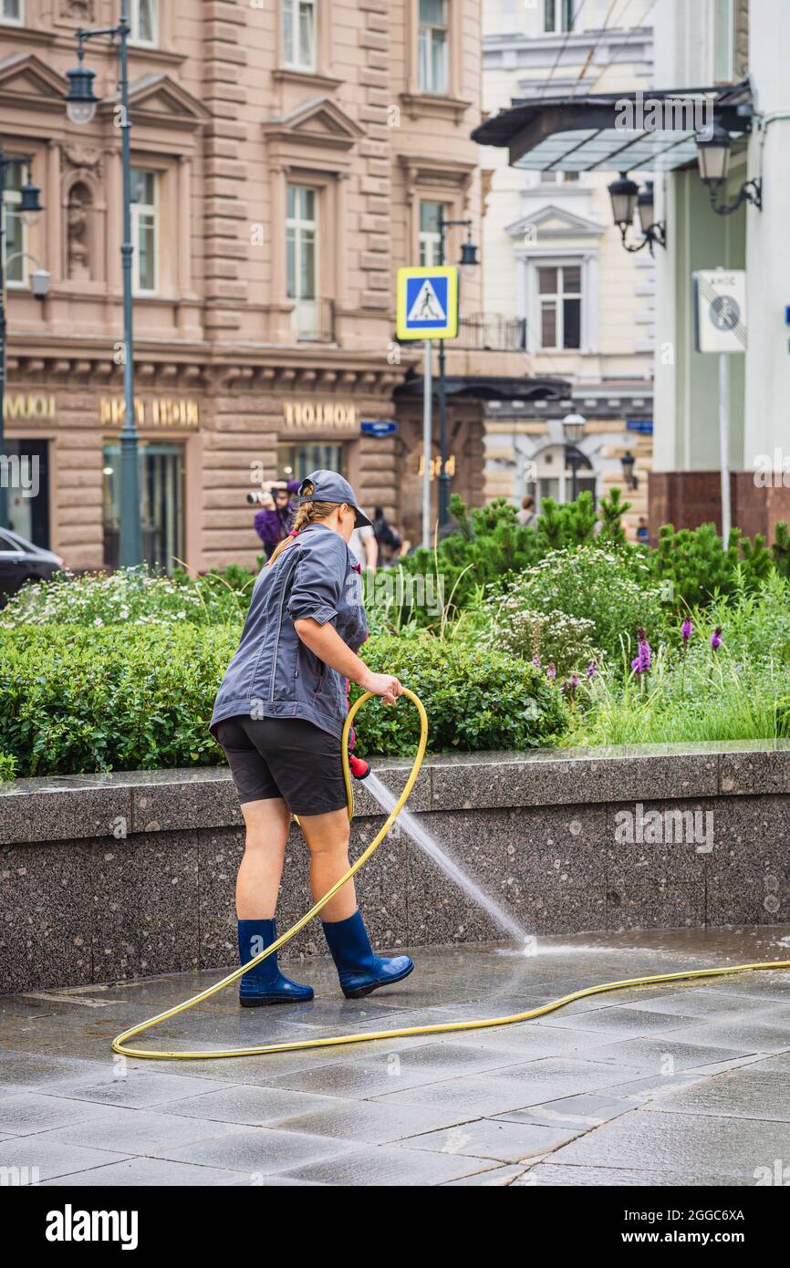 Vue arrière d'une fille méconnue du service de ville arroser des fleurs dans un lit de fleurs et rafraîchir la chaussée par une chaude journée d'été. Les gens ordinaires en public Banque D'Images