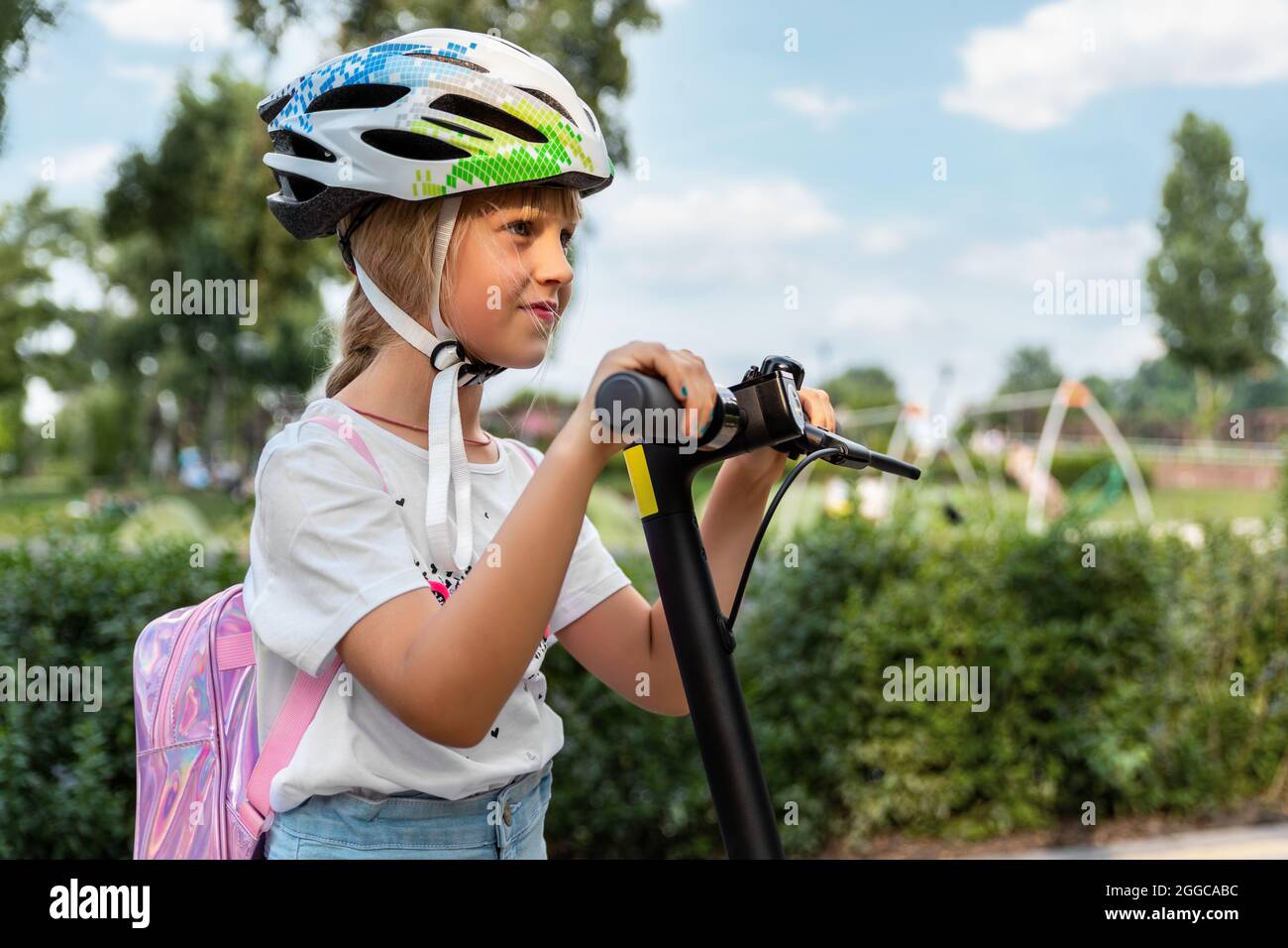 Profil vue portrait de mignon blond petite école caucasienne fille port casque profiter d'avoir plaisir à l'équitation électrique scooter ville parc de rue à l'extérieur sur Banque D'Images