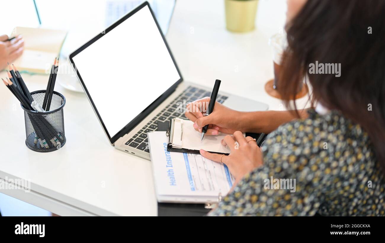 Courtier d'assurance prenant des notes sur ordinateur portable dans des blocs-notes, formulaire d'information d'assurance maladie pour le client sur le bureau, image rognée Banque D'Images