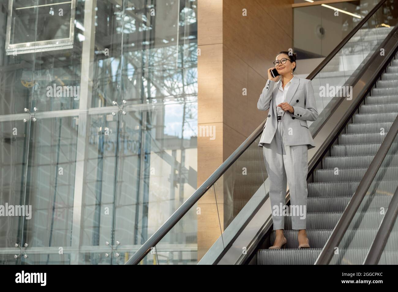 Bonne femme d'affaires en costume gris parlant sur un smartphone tout en descendant sur un escalier roulant Banque D'Images