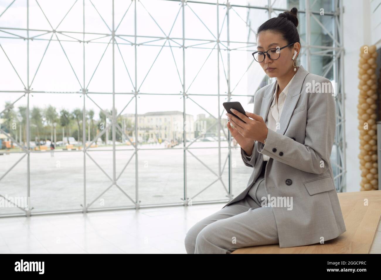 Jeune femme d'affaires sérieuse avec écouteurs regardant la vidéo dans un smartphone tout en étant assise dans un centre d'affaires contemporain Banque D'Images