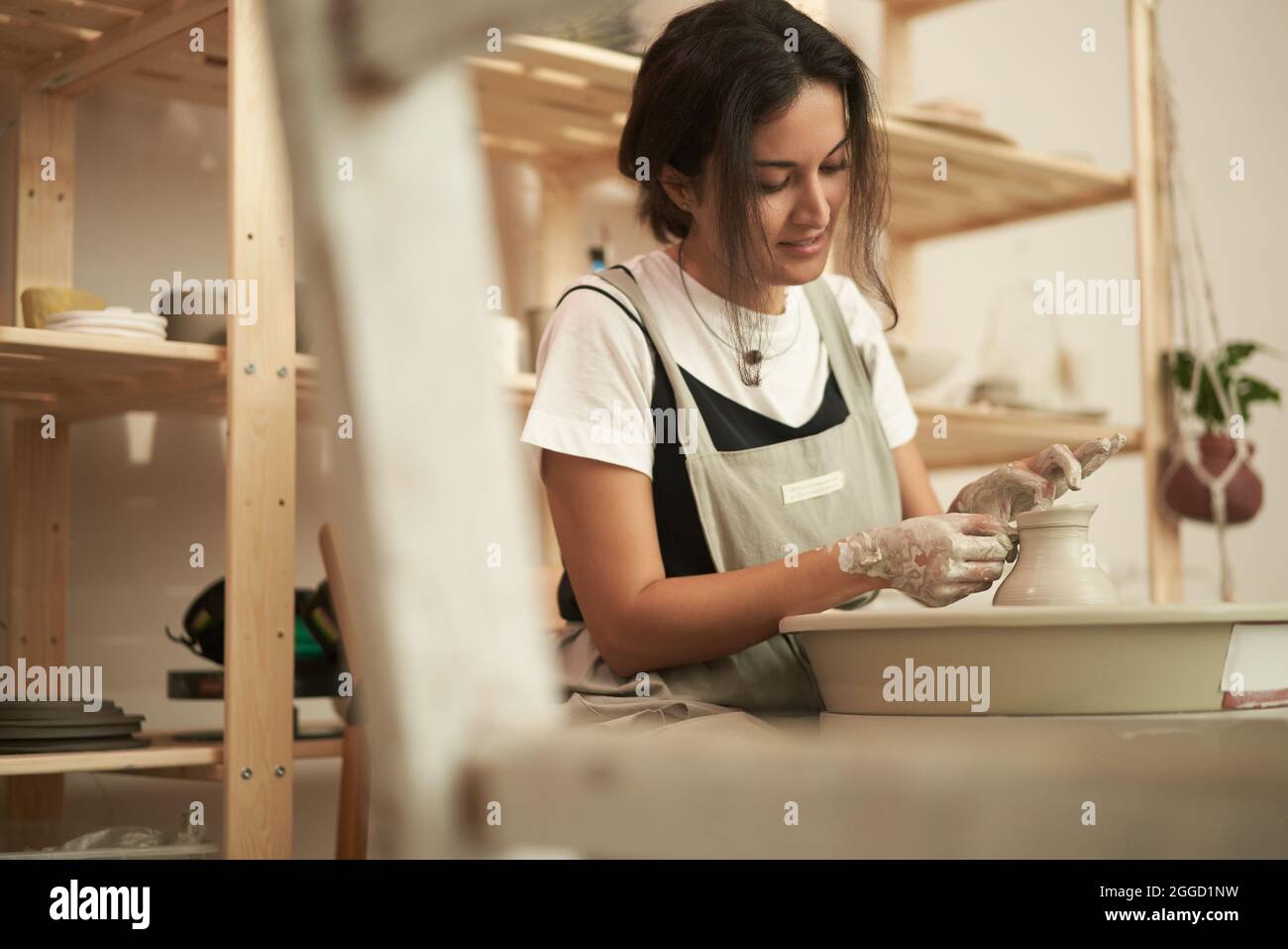 Jeune céramiste féminine qualifiée en tablier sculptant les bords de la marmite à la roue de poterie tout en façonnant la faïence dans un studio d'art créatif Banque D'Images