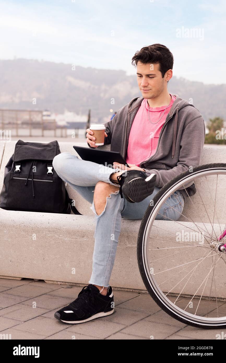 photo verticale d'un jeune homme assis à l'extérieur avec un café à l'aide d'une tablette à côté de son vélo, concept de technologie et de style de vie urbain, espace de copie pour Banque D'Images