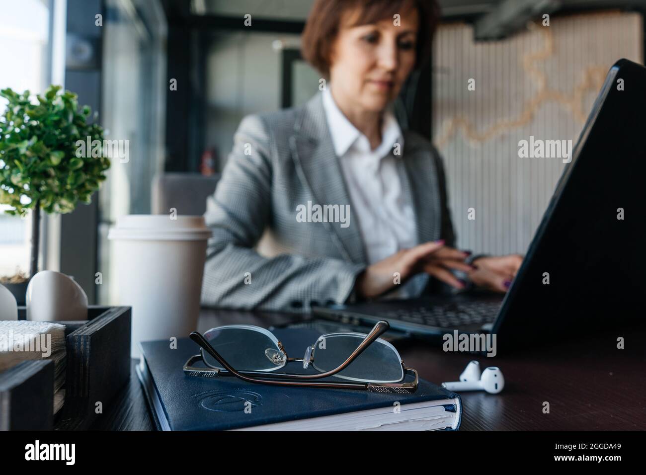Lunettes au premier plan. Femme d'affaires senior dans un blazer blanc travaille dans un café en arrière-plan. Travail à distance. Banque D'Images